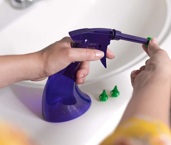 Close-up of a person using a purple spray bottle in a bathroom sink with green nozzles.