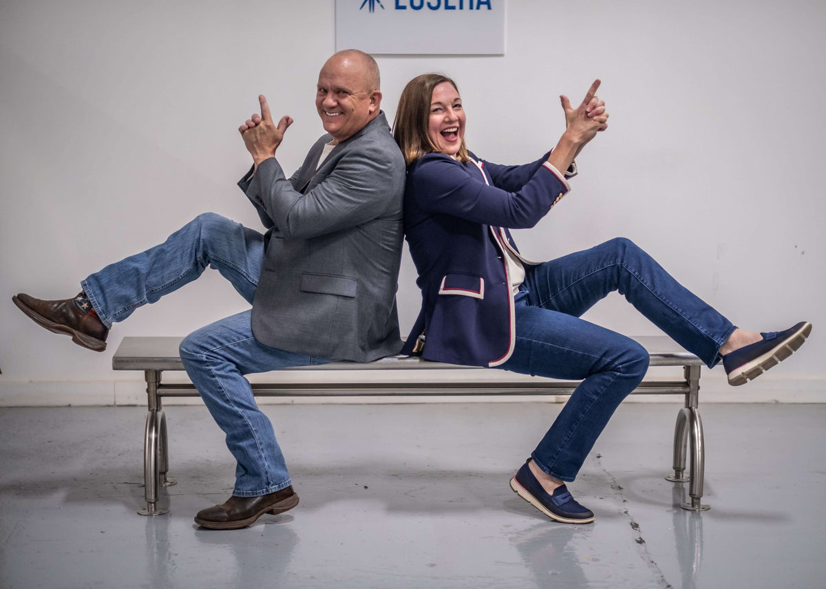 A man and woman sit on a bench and pose with their hands up in the air